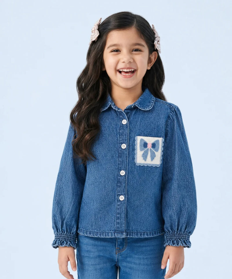 A young girl with long dark hair and pink hair clips is smiling, wearing a blue denim button-up shirt with a decorative bow patch on the chest pocket, paired with blue jeans, against a light blue studio background.