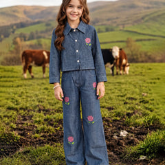 A young girl stands in a grassy field wearing a matching blue denim two-piece outfit with pink floral embroidery. Cows are visible in the background against rolling green hills.