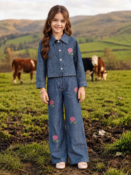 A young girl stands in a grassy field wearing a matching blue denim two-piece outfit with pink floral embroidery. Cows are visible in the background against rolling green hills.