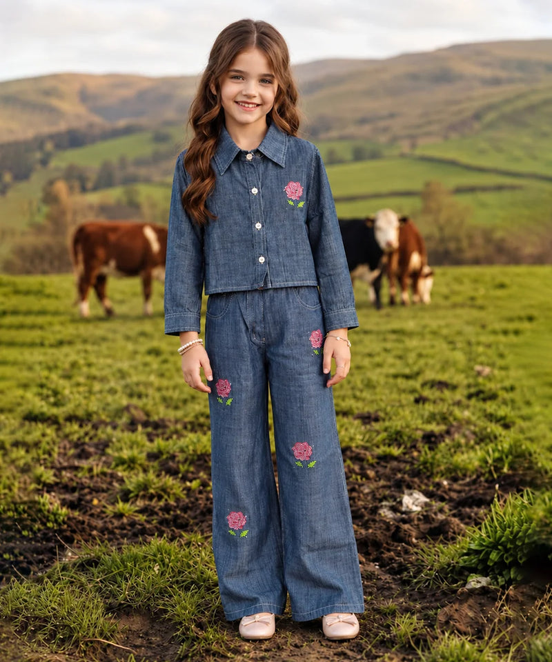 A young girl stands in a grassy field wearing a matching blue denim two-piece outfit with pink floral embroidery. Cows are visible in the background against rolling green hills.