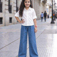 A young girl stands on a city sidewalk wearing a white short-sleeve button-up shirt and wide-leg blue jeans, with her hands on her hips.
