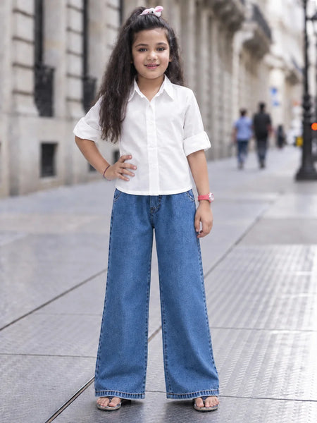 A young girl stands on a city sidewalk wearing a white short-sleeve button-up shirt and wide-leg blue jeans, with her hands on her hips.