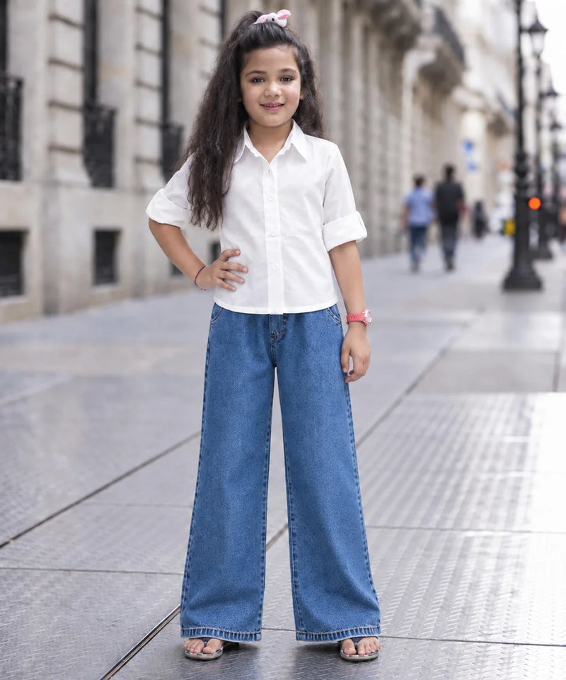 A young girl stands on a city sidewalk wearing a white short-sleeve button-up shirt and wide-leg blue jeans, with her hands on her hips.