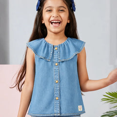 A young girl smiling and laughing while wearing a light blue denim sleeveless top with a ruffled collar and gold-tone buttons, paired with matching blue jeans. She has blue hair bows in her hair, set against a soft gray and pink background with a hint of green foliage.