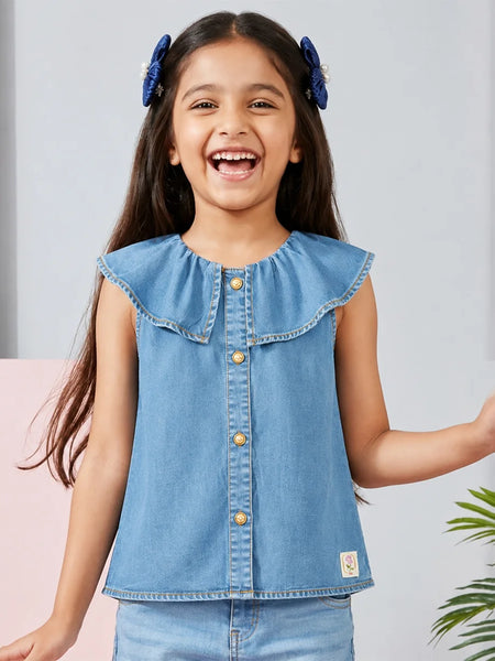 A young girl smiling and laughing while wearing a light blue denim sleeveless top with a ruffled collar and gold-tone buttons, paired with matching blue jeans. She has blue hair bows in her hair, set against a soft gray and pink background with a hint of green foliage.