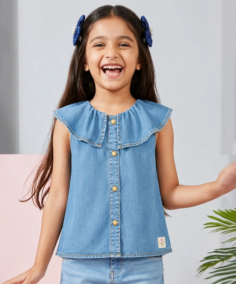 A young girl smiling and laughing while wearing a light blue denim sleeveless top with a ruffled collar and gold-tone buttons, paired with matching blue jeans. She has blue hair bows in her hair, set against a soft gray and pink background with a hint of green foliage.
