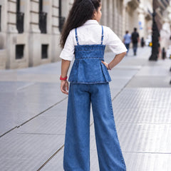A young girl seen from behind wearing a blue denim overall-style top with wide-leg pants over a white short-sleeve shirt, standing on a city sidewalk.