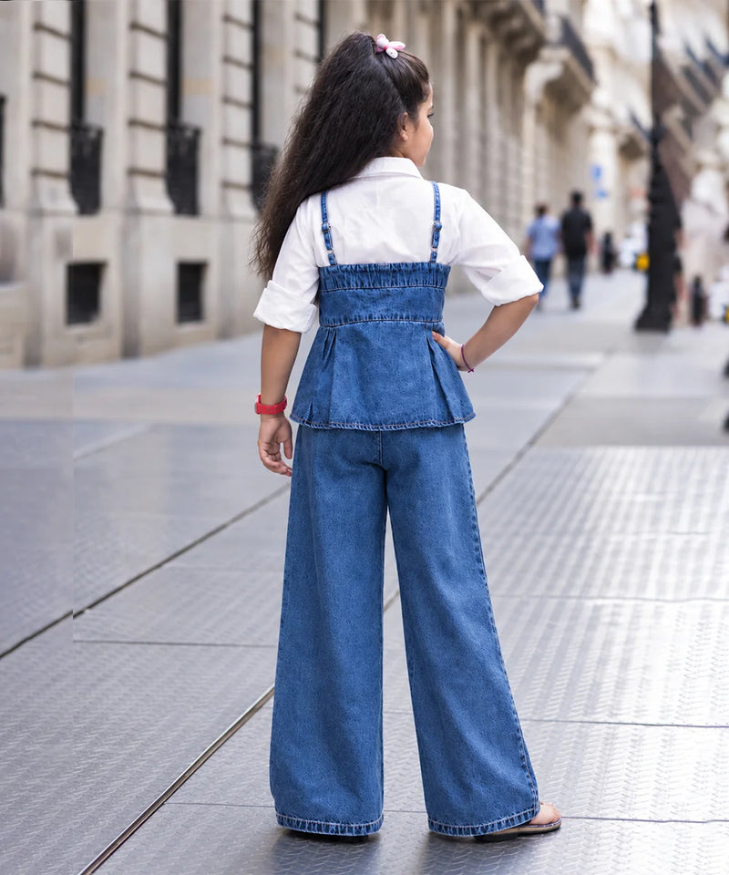 A young girl seen from behind wearing a blue denim overall-style top with wide-leg pants over a white short-sleeve shirt, standing on a city sidewalk.