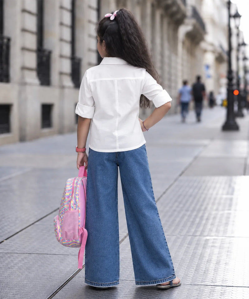 A young girl seen from behind wearing a white short-sleeve button-up shirt and wide-leg blue jeans, standing on a city sidewalk and holding a pink backpack.