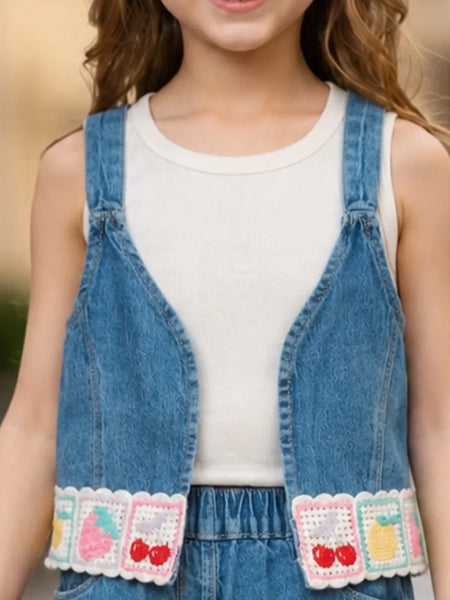 A child wearing a blue denim overall-style vest over a white tank top, with decorative crocheted fruit and fruit-with-cherries patches along the bottom front. The photo is cropped from the lower face to the hips against a softly blurred outdoor background.
