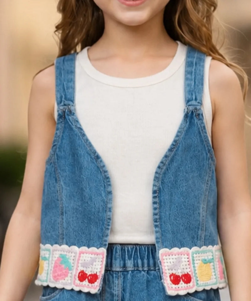 A child wearing a blue denim overall-style vest over a white tank top, with decorative crocheted fruit and fruit-with-cherries patches along the bottom front. The photo is cropped from the lower face to the hips against a softly blurred outdoor background.