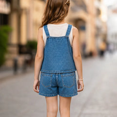 A young girl viewed from behind wearing a sleeveless blue denim overall romper with matching shorts over a white tank top, standing on a blurred cobblestone street.