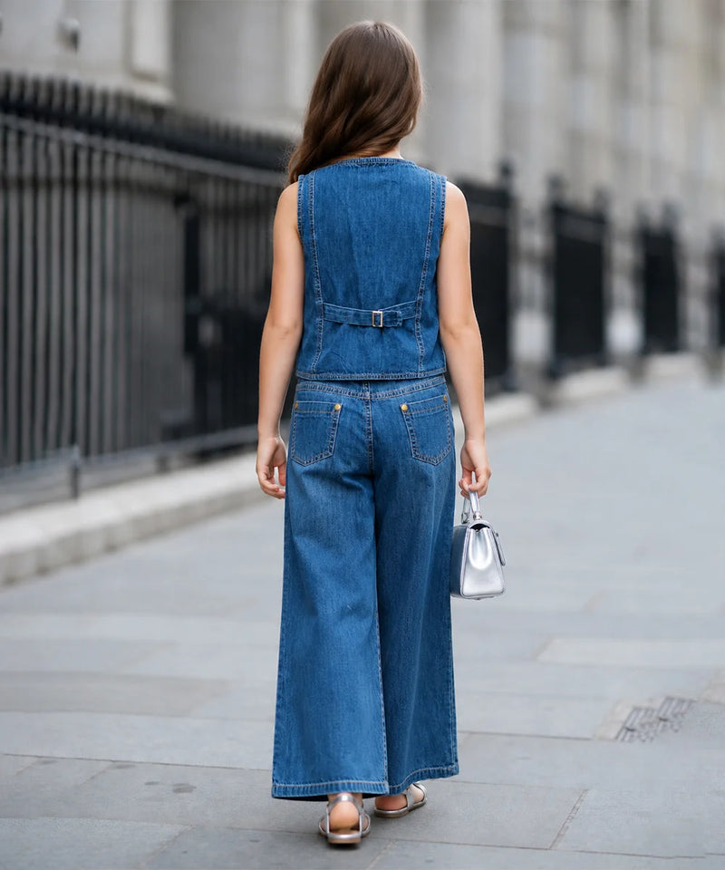 A woman viewed from behind wearing a sleeveless blue denim jumpsuit with wide-leg pants and a small silver handbag on a city sidewalk.