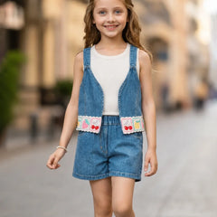 A young girl viewed from behind wearing a sleeveless blue denim overall romper with matching shorts over a white tank top, standing on a blurred cobblestone street.