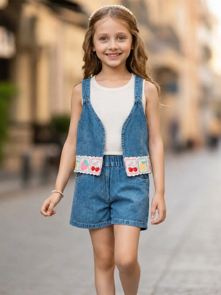 A young girl viewed from behind wearing a sleeveless blue denim overall romper with matching shorts over a white tank top, standing on a blurred cobblestone street.