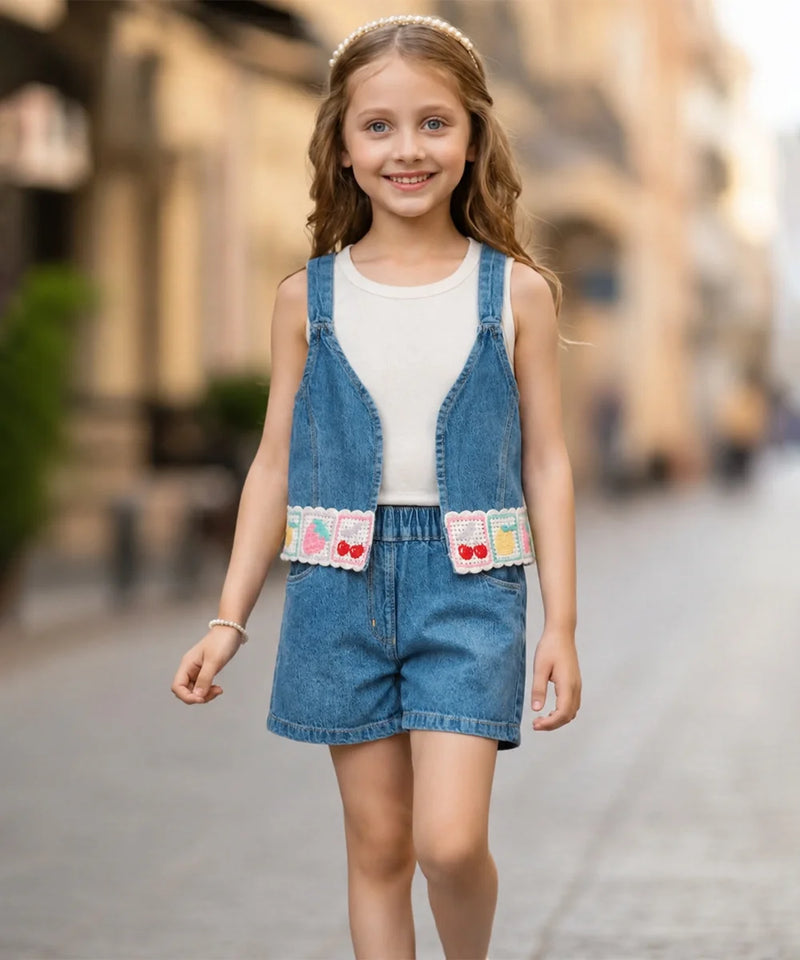 A young girl viewed from behind wearing a sleeveless blue denim overall romper with matching shorts over a white tank top, standing on a blurred cobblestone street.