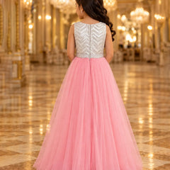 A young girl viewed from behind wearing a formal ball gown with a silver chevron-embellished sleeveless back bodice and a voluminous pink tulle skirt, standing in a grand, warmly lit hall with chandeliers.