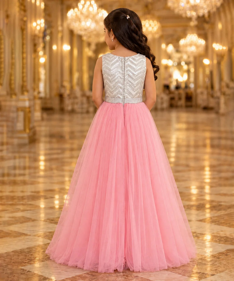 A young girl viewed from behind wearing a formal ball gown with a silver chevron-embellished sleeveless back bodice and a voluminous pink tulle skirt, standing in a grand, warmly lit hall with chandeliers.