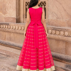 A woman viewed from behind wearing a bright pink sleeveless Anarkali gown with gold stripes and a gold border, standing in front of a beige stone building.