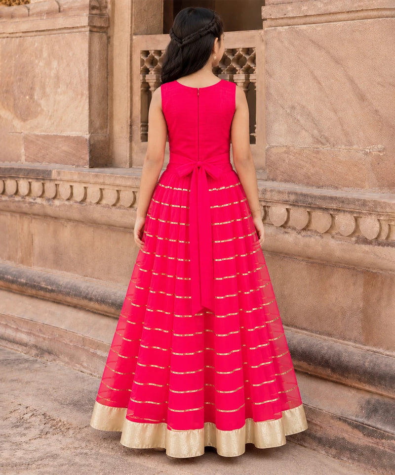 A woman viewed from behind wearing a bright pink sleeveless Anarkali gown with gold stripes and a gold border, standing in front of a beige stone building.