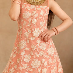 A young girl modeling a peach-colored sleeveless ethnic gown with cream floral embroidery and a draped asymmetrical overlay, set against a warm beige architectural background with a large clay pot.