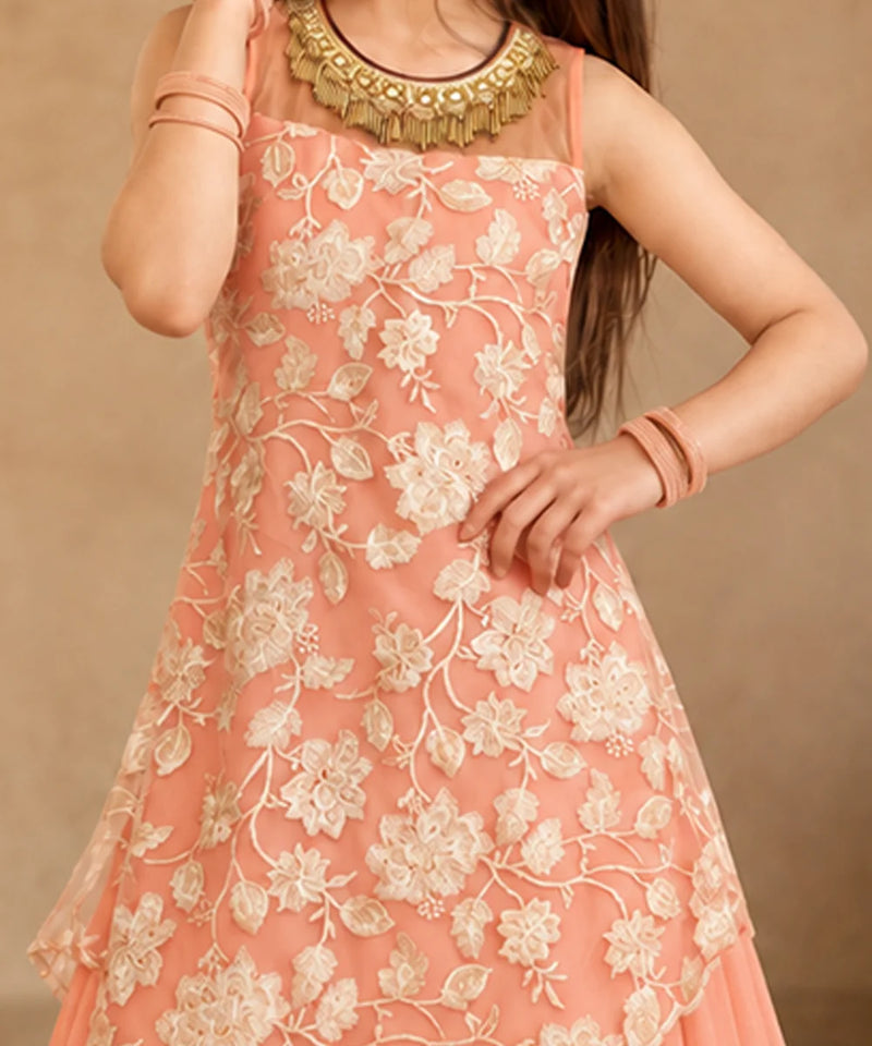 A young girl modeling a peach-colored sleeveless ethnic gown with cream floral embroidery and a draped asymmetrical overlay, set against a warm beige architectural background with a large clay pot.