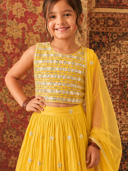 Young girl in a yellow traditional outfit with sequins against a patterned wall.