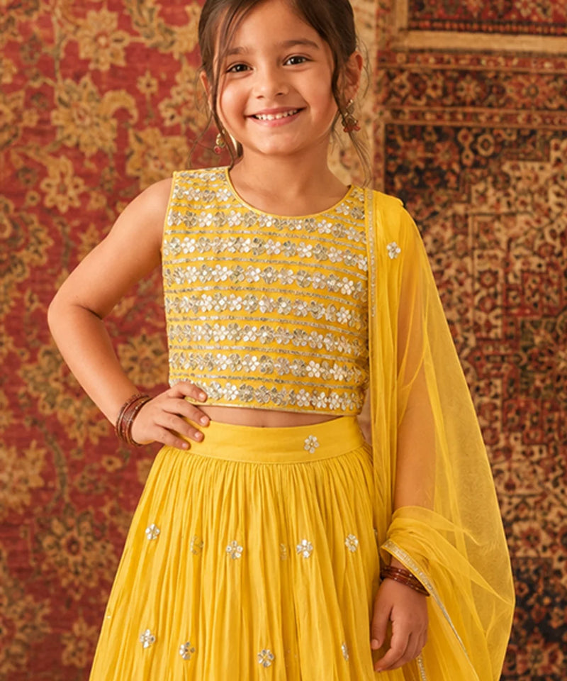Young girl in a yellow traditional outfit with sequins against a patterned wall.
