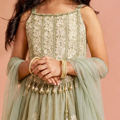 A close-up of a child wearing a sage green embroidered traditional dress with a matching sheer dupatta and gold bangles, posed against a solid peach-pink background.