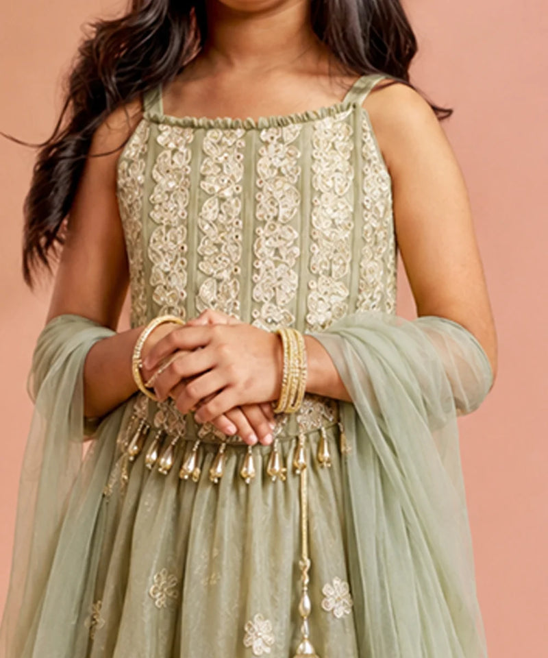 A close-up of a child wearing a sage green embroidered traditional dress with a matching sheer dupatta and gold bangles, posed against a solid peach-pink background.