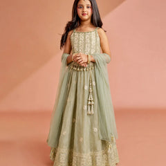 A young girl modeling a sage green Anarkali gown with intricate gold embroidery, a matching sheer dupatta, and a flared skirt, set against a warm peach studio backdrop.