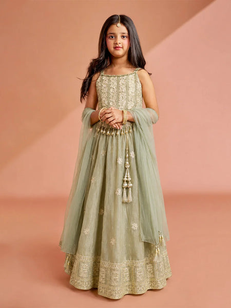 A young girl modeling a sage green Anarkali gown with intricate gold embroidery, a matching sheer dupatta, and a flared skirt, set against a warm peach studio backdrop.