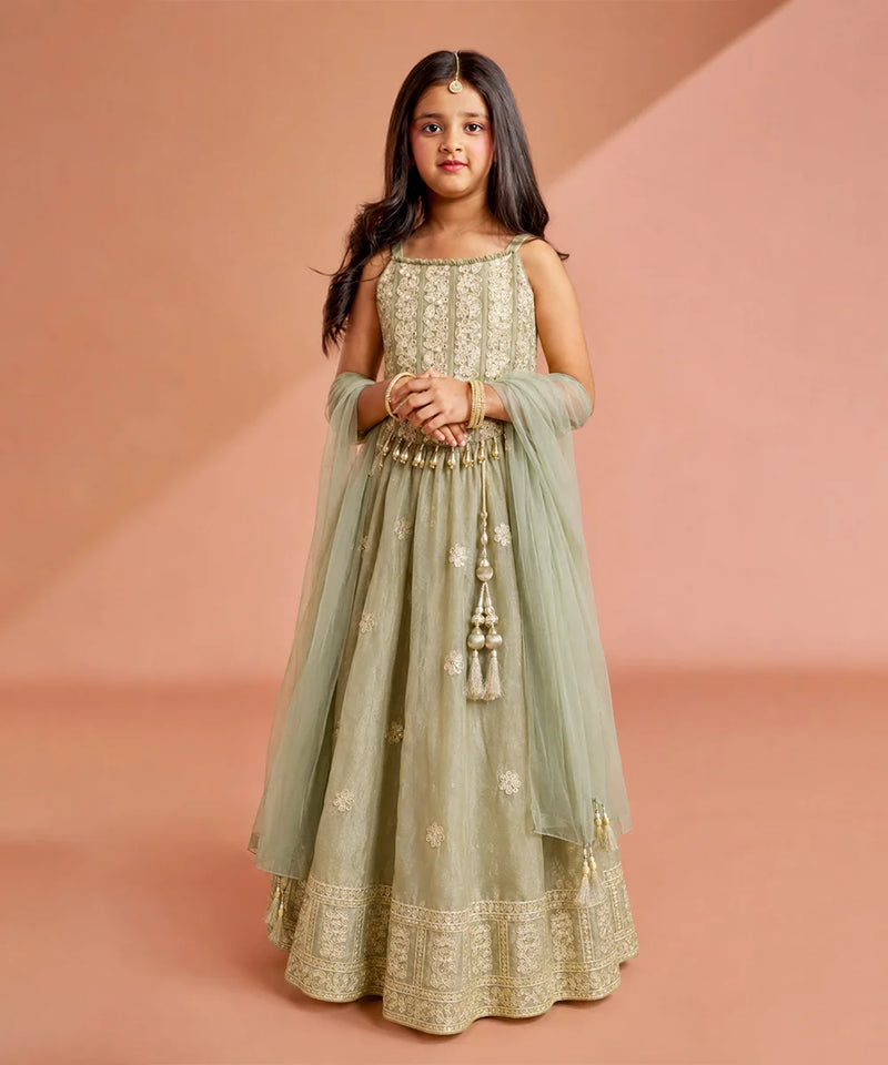 A young girl modeling a sage green Anarkali gown with intricate gold embroidery, a matching sheer dupatta, and a flared skirt, set against a warm peach studio backdrop.
