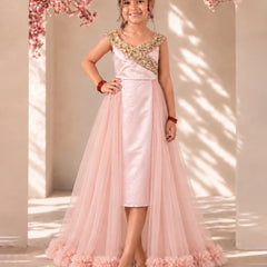 A young girl stands in a sunlit indoor setting wearing a formal light pink dress with a satin bodice, gold embroidered yoke, and a full tulle skirt with ruffled trim.