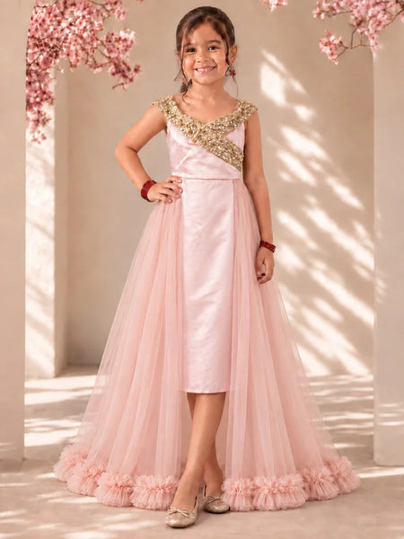 A young girl stands in a sunlit indoor setting wearing a formal light pink dress with a satin bodice, gold embroidered yoke, and a full tulle skirt with ruffled trim.