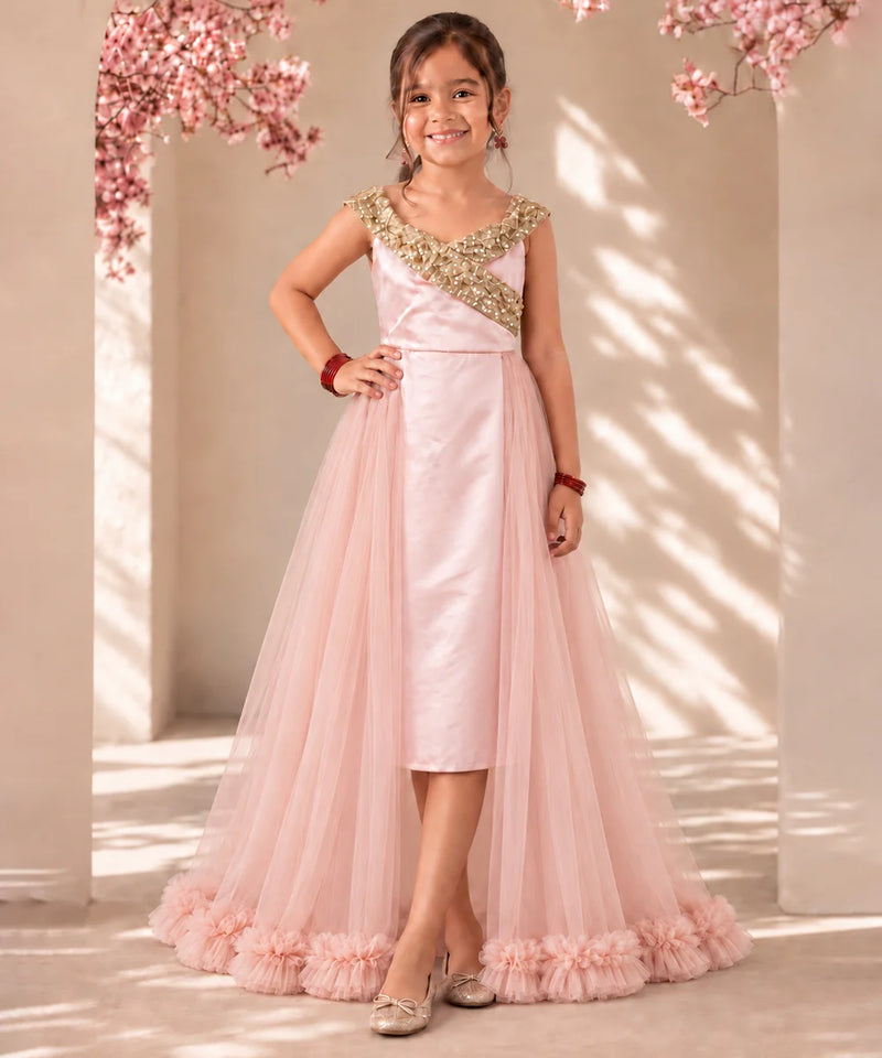 A young girl stands in a sunlit indoor setting wearing a formal light pink dress with a satin bodice, gold embroidered yoke, and a full tulle skirt with ruffled trim.
