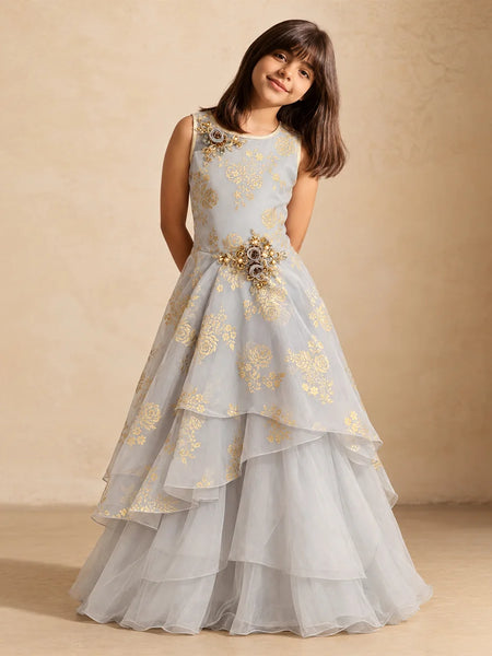 A young girl wearing a sleeveless light blue-gray gown with gold floral embroidery and a jeweled waist detail, standing against a warm beige textured backdrop.