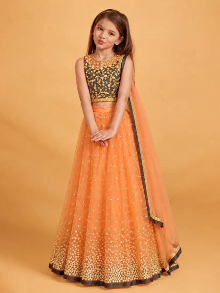 A young girl modeling a traditional Indian ethnic wear set consisting of an orange nette lehenga skirt with gold foil work, a black embroidered choli, and a matching orange dupatta, set against a solid warm beige background.