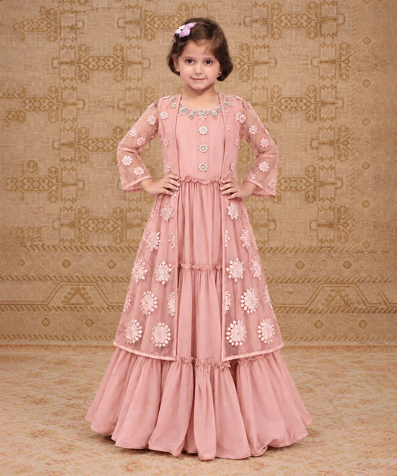 A pink traditional Indian dress with intricate floral embroidery and beadwork, shown on a model against a patterned beige background.