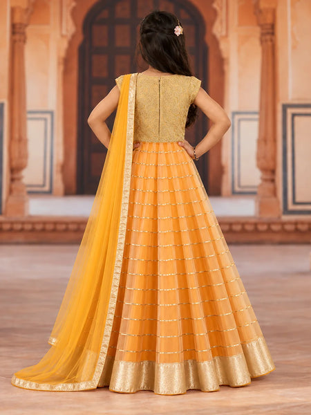 A woman viewed from behind wearing a traditional Indian outfit consisting of a gold embroidered sleeveless kurta, a pleated yellow-orange skirt, and a matching yellow dupatta, set against a warm architectural background.