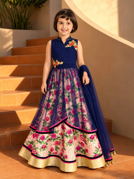 A young girl stands on terracotta steps wearing a sleeveless navy blue and floral pink traditional dress with gold embroidery and a matching navy dupatta.