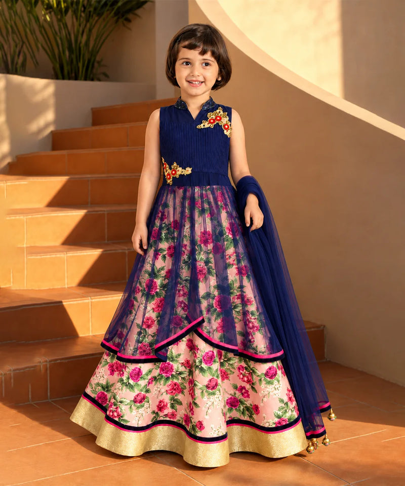A young girl stands on terracotta steps wearing a sleeveless navy blue and floral pink traditional dress with gold embroidery and a matching navy dupatta.