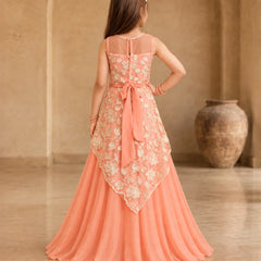 A young girl viewed from behind wearing a peach and beige floral lace formal gown with a large bow at the waist, standing in a warm-toned indoor setting with a large vase in the background.