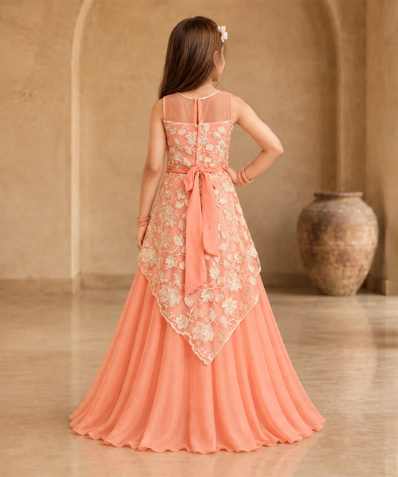 A young girl viewed from behind wearing a peach and beige floral lace formal gown with a large bow at the waist, standing in a warm-toned indoor setting with a large vase in the background.