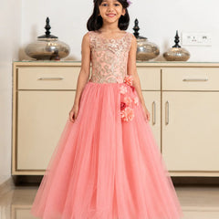 A young girl stands in a room wearing a formal sleeveless gown with a beige floral lace bodice and a voluminous pink tulle skirt decorated with 3D floral appliqués.