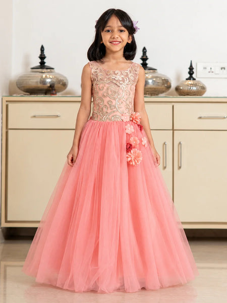 A young girl stands in a room wearing a formal sleeveless gown with a beige floral lace bodice and a voluminous pink tulle skirt decorated with 3D floral appliqués.