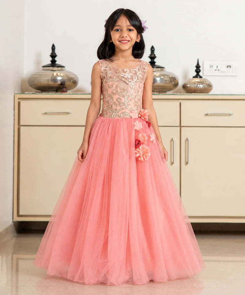 A young girl stands in a room wearing a formal sleeveless gown with a beige floral lace bodice and a voluminous pink tulle skirt decorated with 3D floral appliqués.