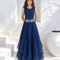 A woman stands in a white architectural setting wearing a sleeveless navy blue maxi dress with a floral embroidered waistband. The dress features a pleated tulle skirt with a layered high-low hem.
