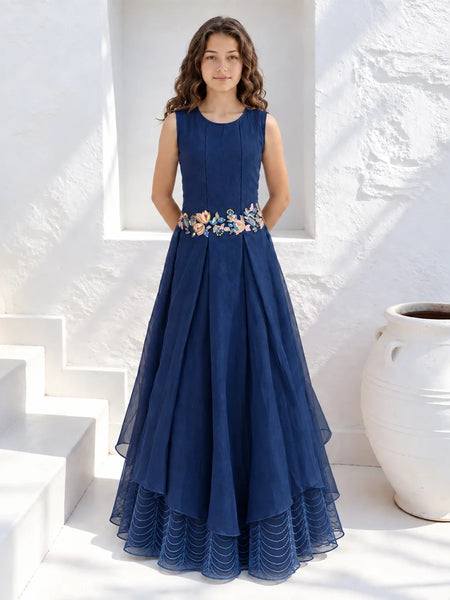 A woman stands in a white architectural setting wearing a sleeveless navy blue maxi dress with a floral embroidered waistband. The dress features a pleated tulle skirt with a layered high-low hem.