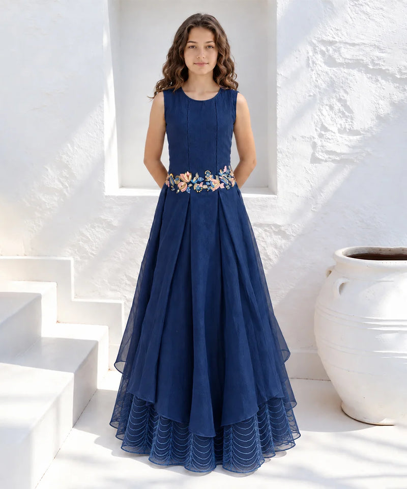 A woman stands in a white architectural setting wearing a sleeveless navy blue maxi dress with a floral embroidered waistband. The dress features a pleated tulle skirt with a layered high-low hem.